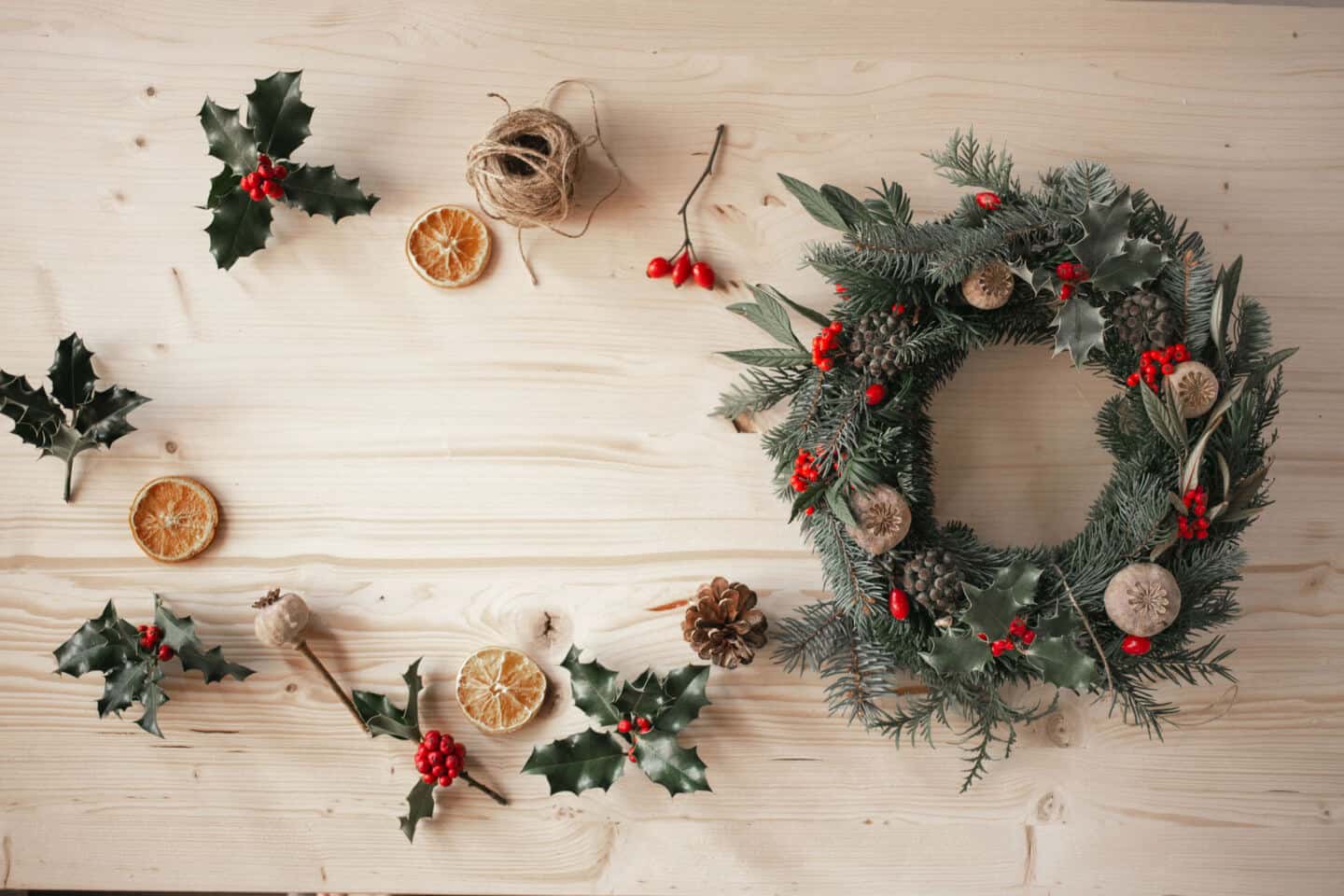 A Christmas wreath on a wooden table with string, holy, pine cones and dry slices of orange spread across the table