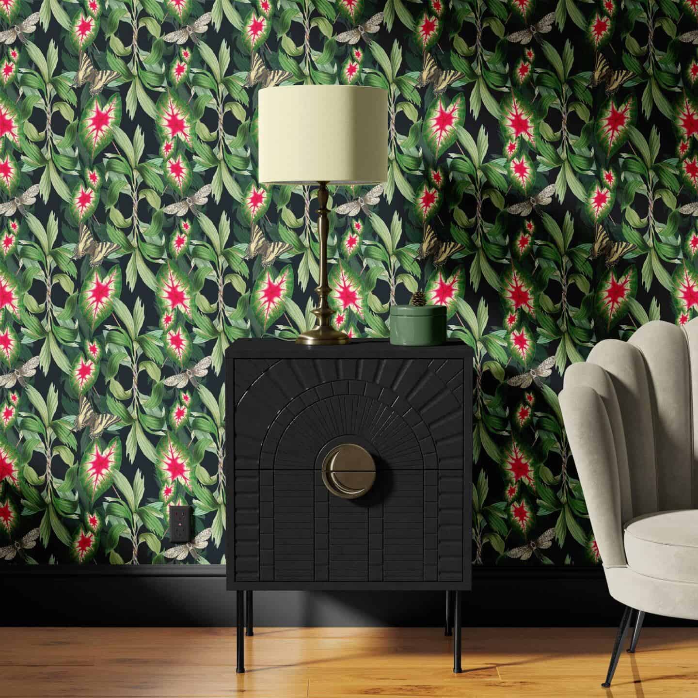 A living room featuring a grey scalloped chair and a small black cabinet with table lamp on it. Black, green and pink botanical wallpaper is on the wall behind.