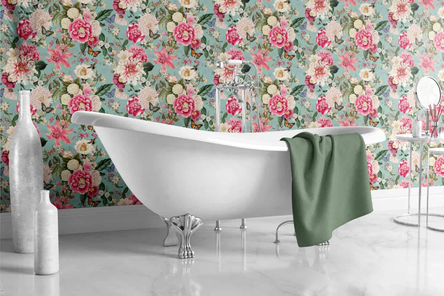 A white free-standing bathtub on a marble floor with a green towel hanging over the side. A beautiful pink and blue floral wallpaper is on the walls