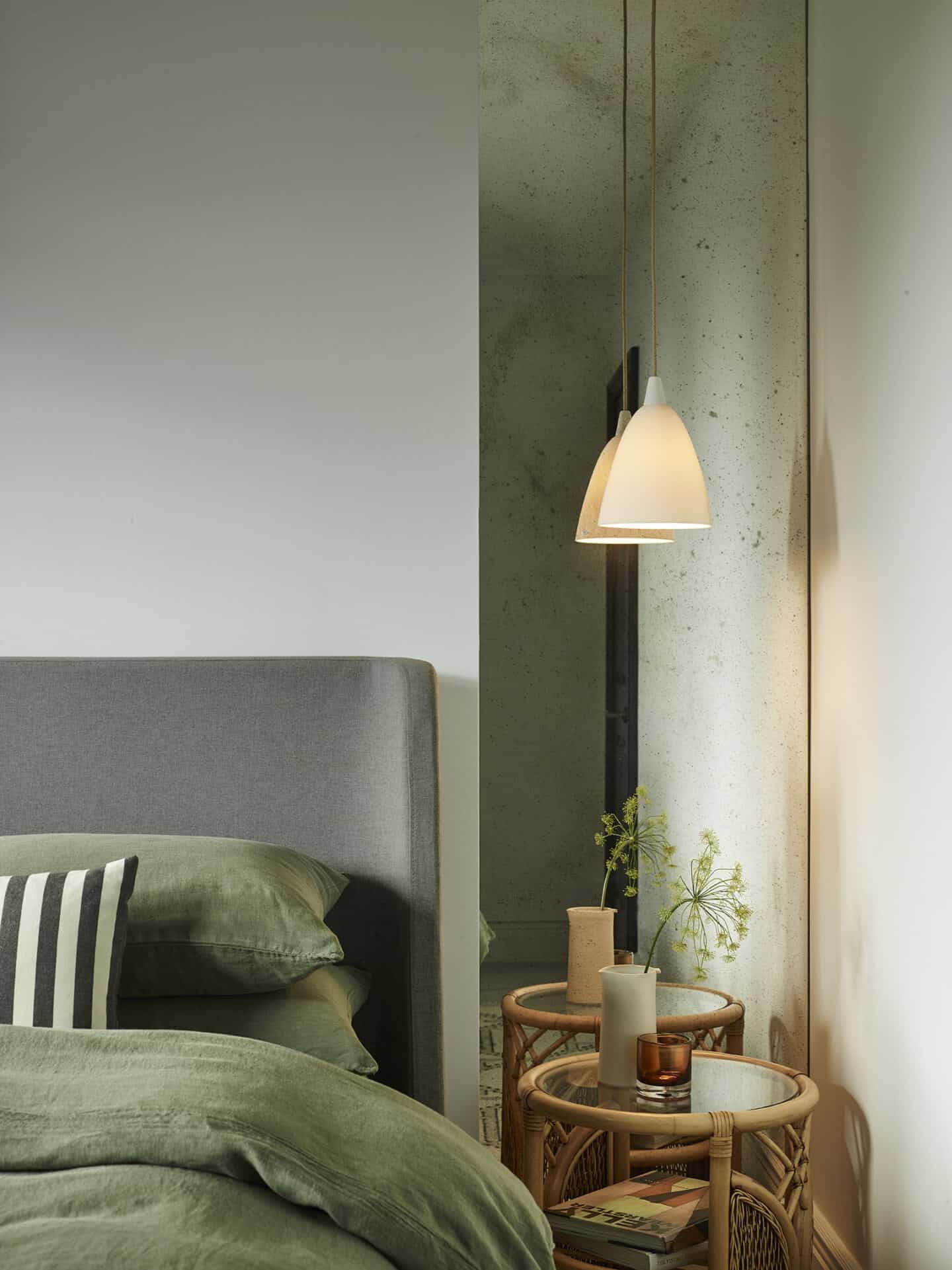 A pendant light hangs down in front of a mirror and above a rattan side table in a green bedroom.