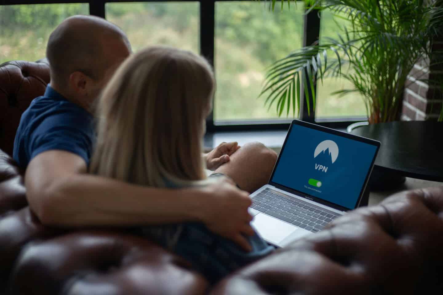 A man and a woman sit together on a brown leather chesterfield sofa looking at a laptop. He has her arm around her. They are using a VPN for added home security