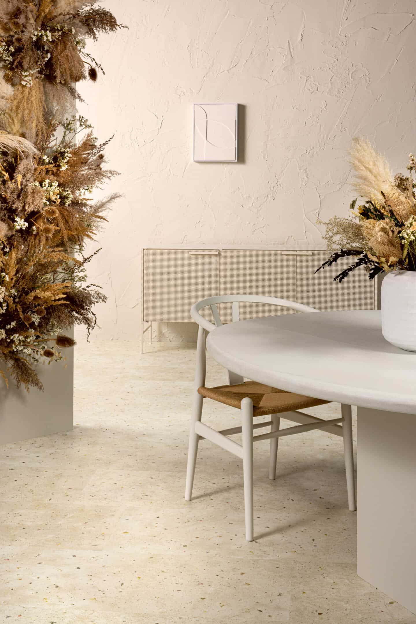 A rattan sideboard on a nature-inspired laminate flooring. There is a dining table with a vase of dried flowers on it in the foreground. The wall finish is very rustic and uneven. Flooring is Botanic Concrete from Quickstep