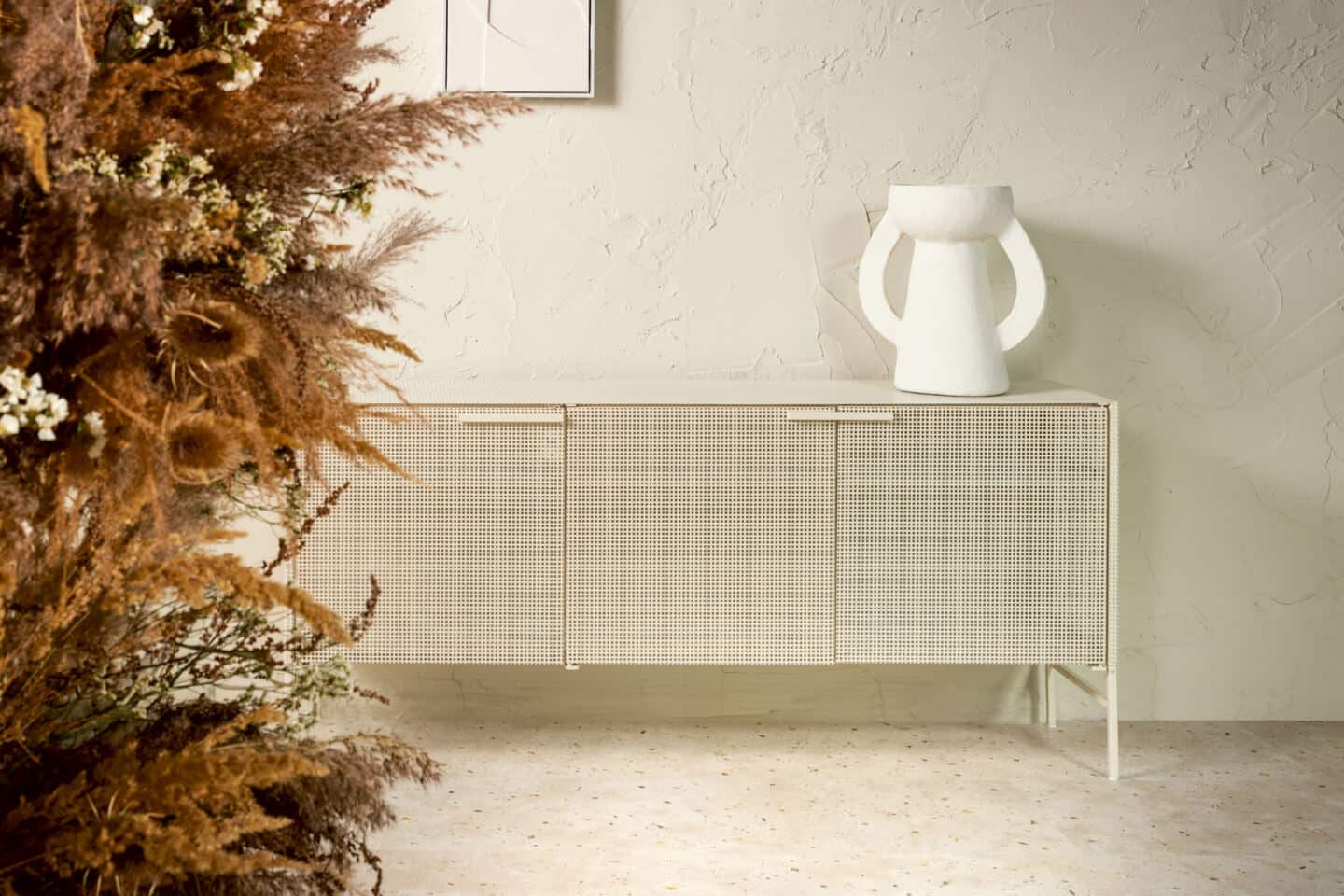 A rattan sideboard on a nature-inspired laminate flooring. There are dried flowers in the foreground and an organic vase on the sideboard. The wall finish is very rustic and uneven. Flooring is Botanic Concrete from Quickstep