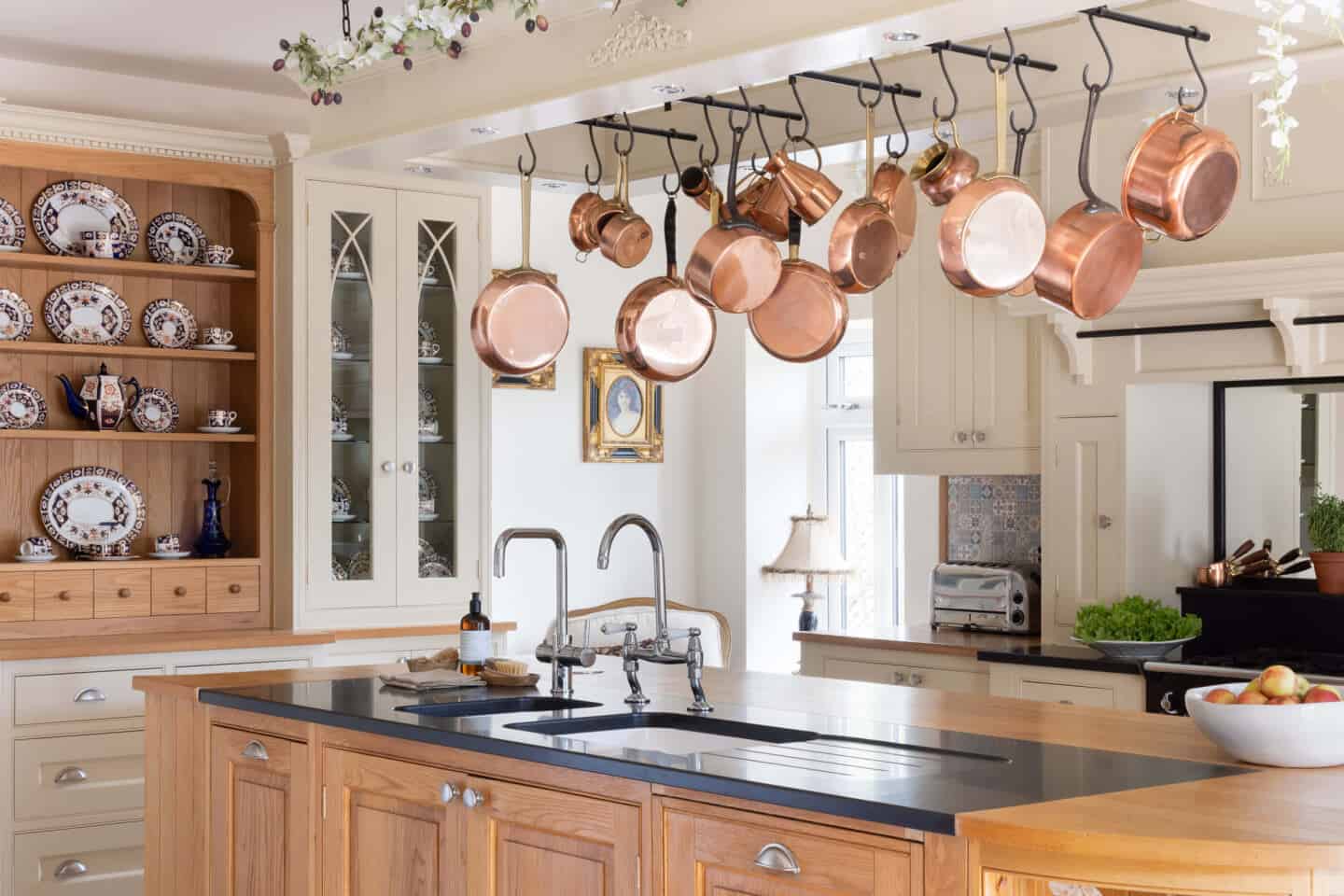 A traditional country kitchen in cream with a central island. Above the island hangs a pot rack full of copper pots