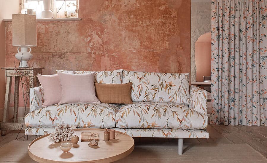 Made-to-Measure Curtains & Blinds using Romo fabric. A sofa in botanical print sits in front of a distressed terracotta wall with long curtains to the right and a round wooden coffee table in front.