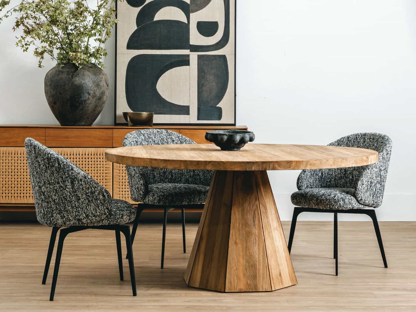 A large round wooden dining table and upholstered dining chairs. A sideboard with woven rattan doors in the background with a large vase on it and a geometric abstract art print