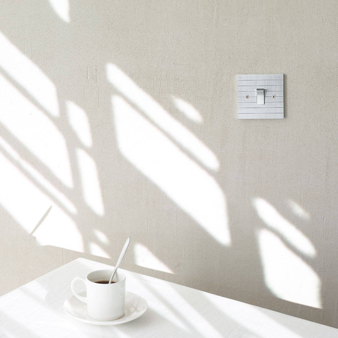 Designer light switches by Kelly Hoppen for Focus SB. A silver light switch on a grey wall above a white table with a cup of black coffee on it.