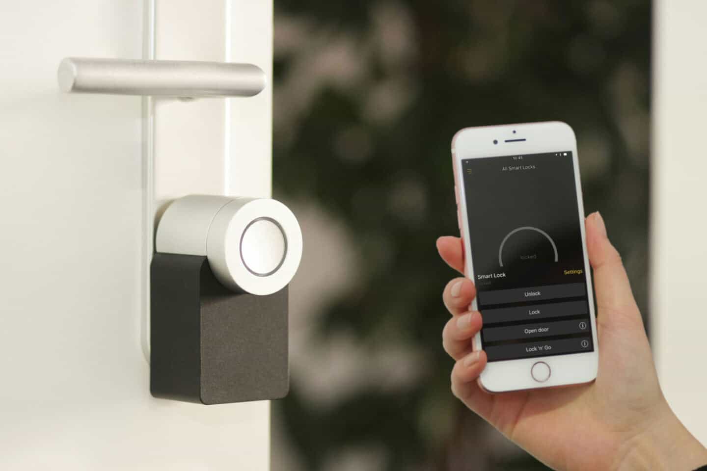 A woman holds a mobile phone with a home security app on it next to a  smart lock on a door