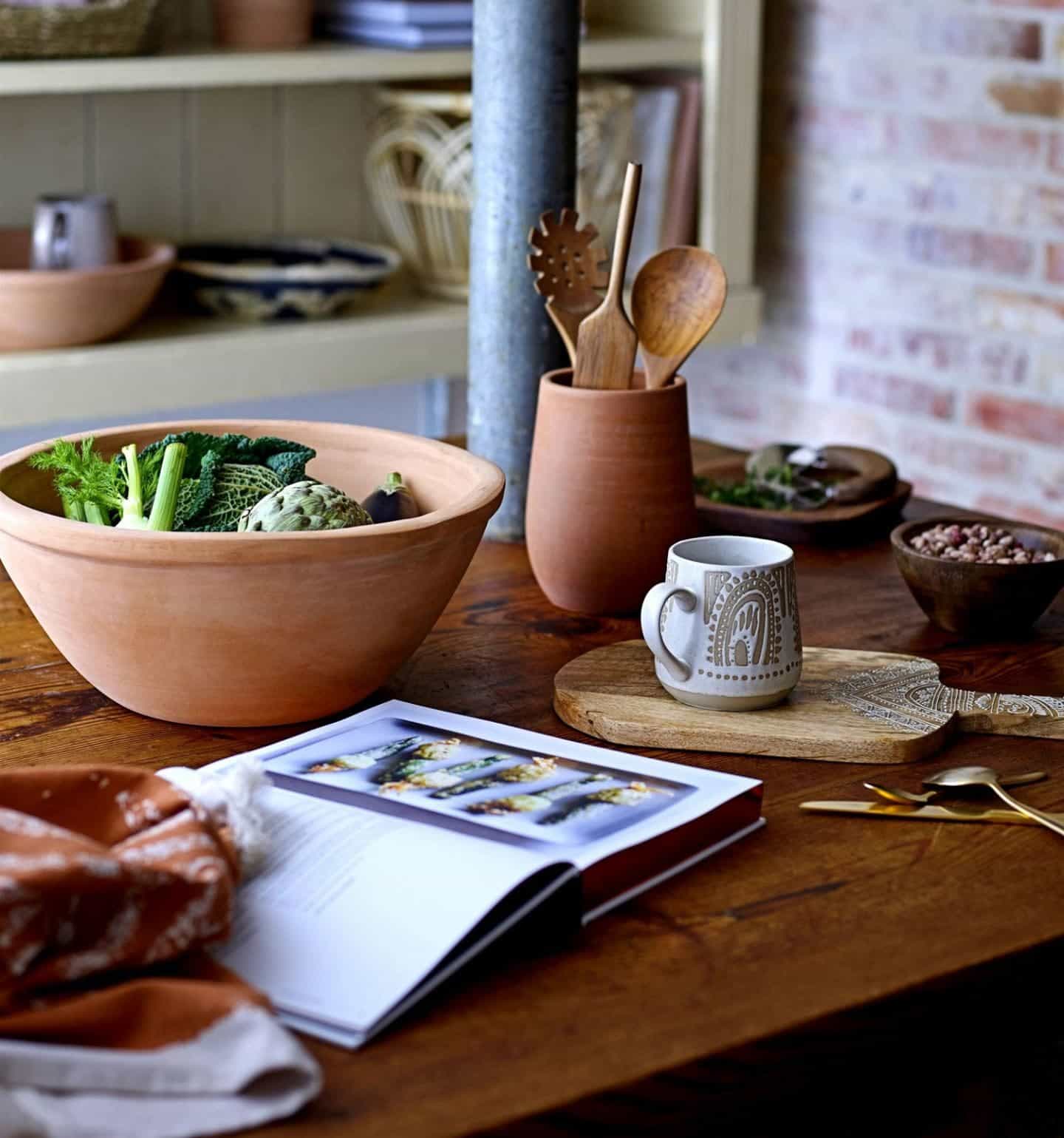 Shekåbba The Danish Home stocks a range of Danish homewares. An earthenware bowl of vegetables on a wooden tabletop next to a pot of wooden utensils a mug of tea and a cookbook.