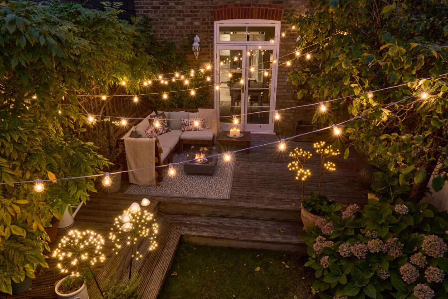 A back garden with cosy seating area lit up with outdoor festoon lights and other solar powered garden lights