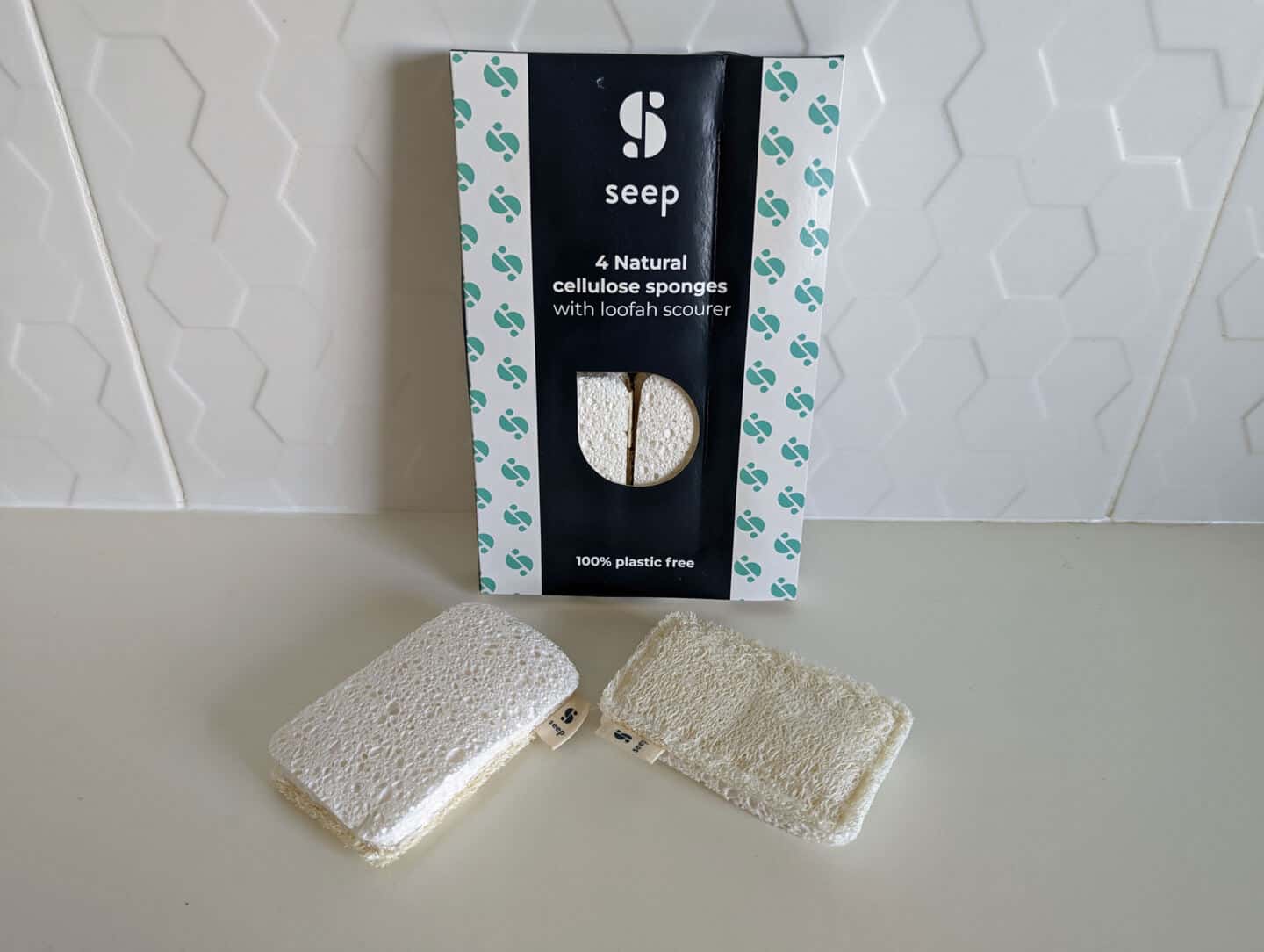 A packet of natural cellulose sponges with loofah scourer on a white work top with white tiles behind.