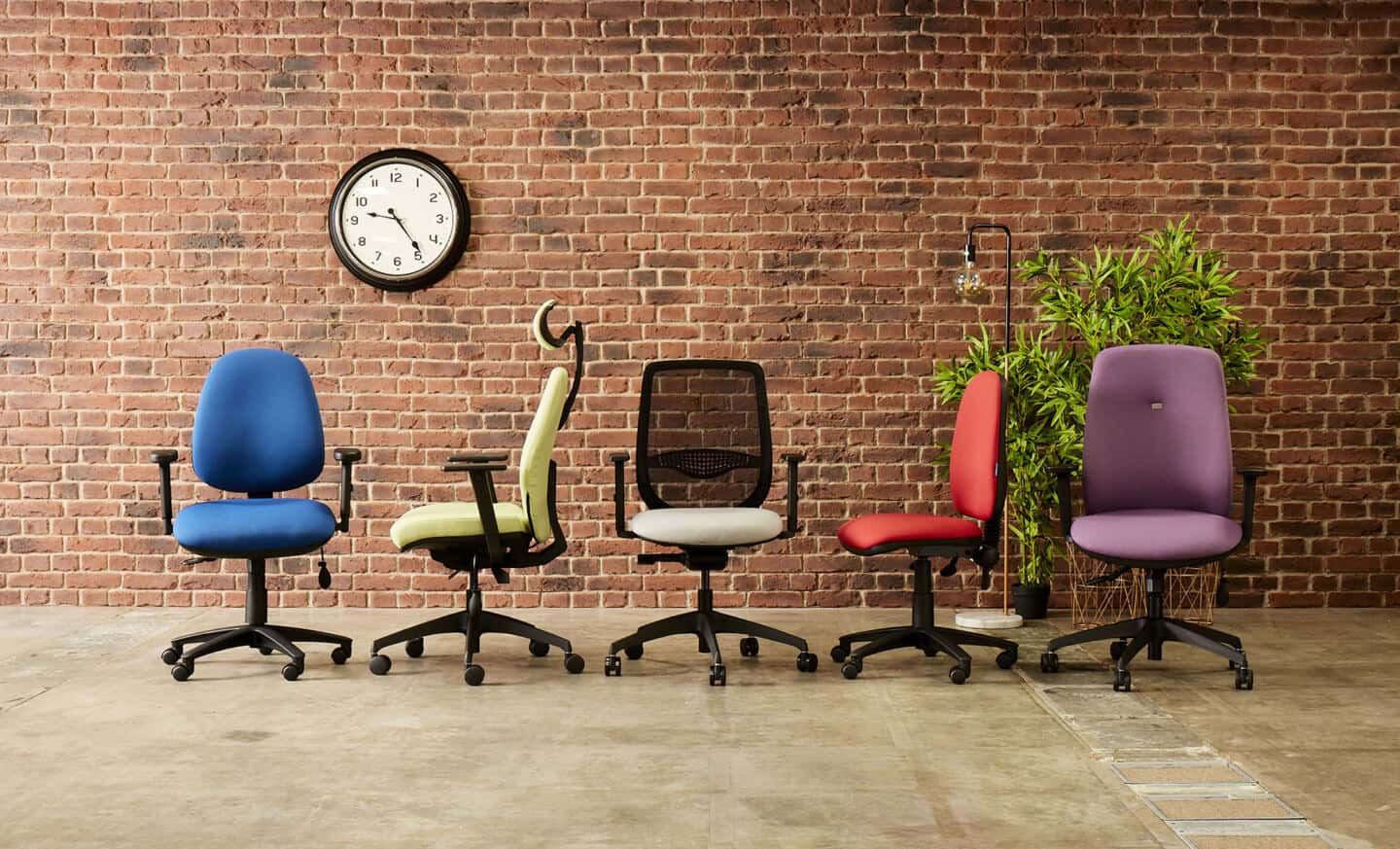 A group of 5 ergonomic office chairs in front of an exposed brick wall with a clock and a plant behind