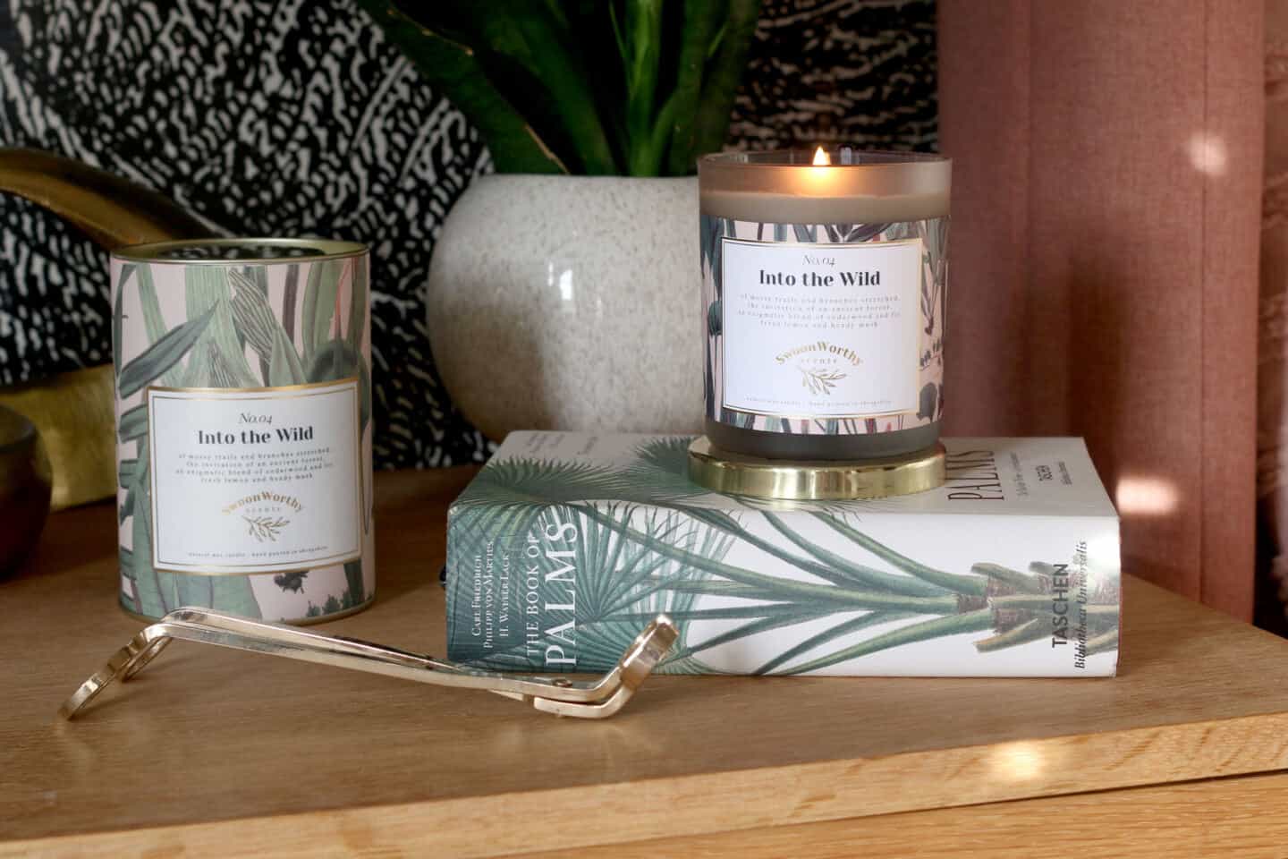 Into the Wild luxury eco-conscious scented candles by Swoon Worthy Scents sat atop a book on a sideboard with a plant behind and a wick trimmer in front.