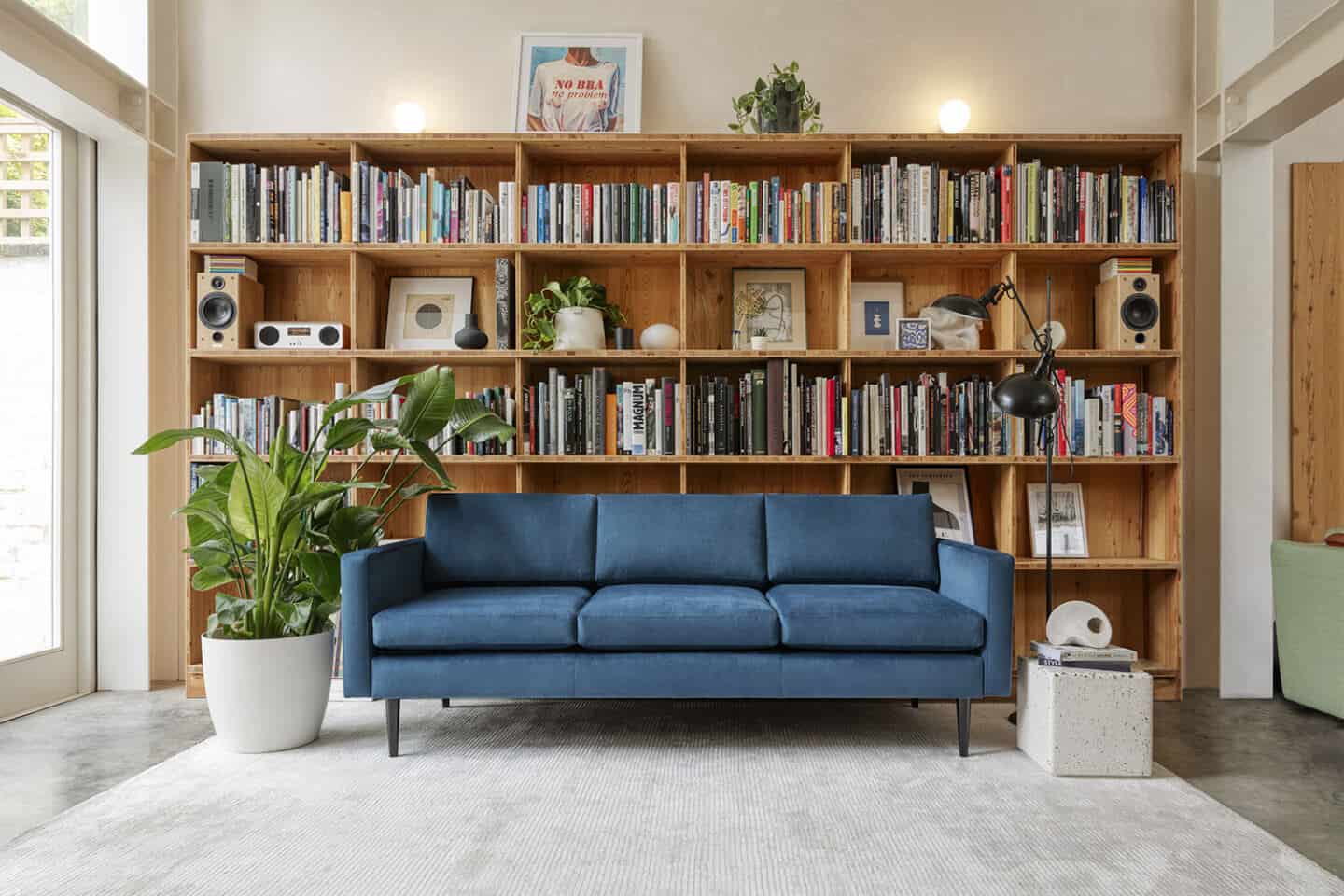 A blue sofa from Swyft Home on a grey rug, positioned next to a window and in front of a large wooden bookcase