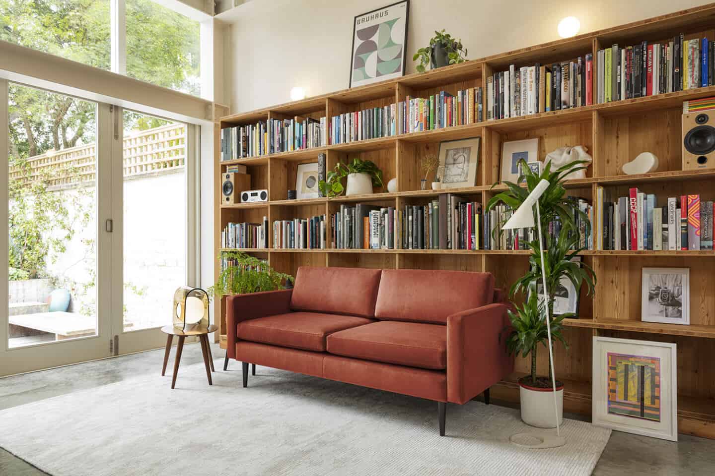 A red sofa from Swyft Home on a grey rug, positioned next to a window and in front of a large wooden bookcase