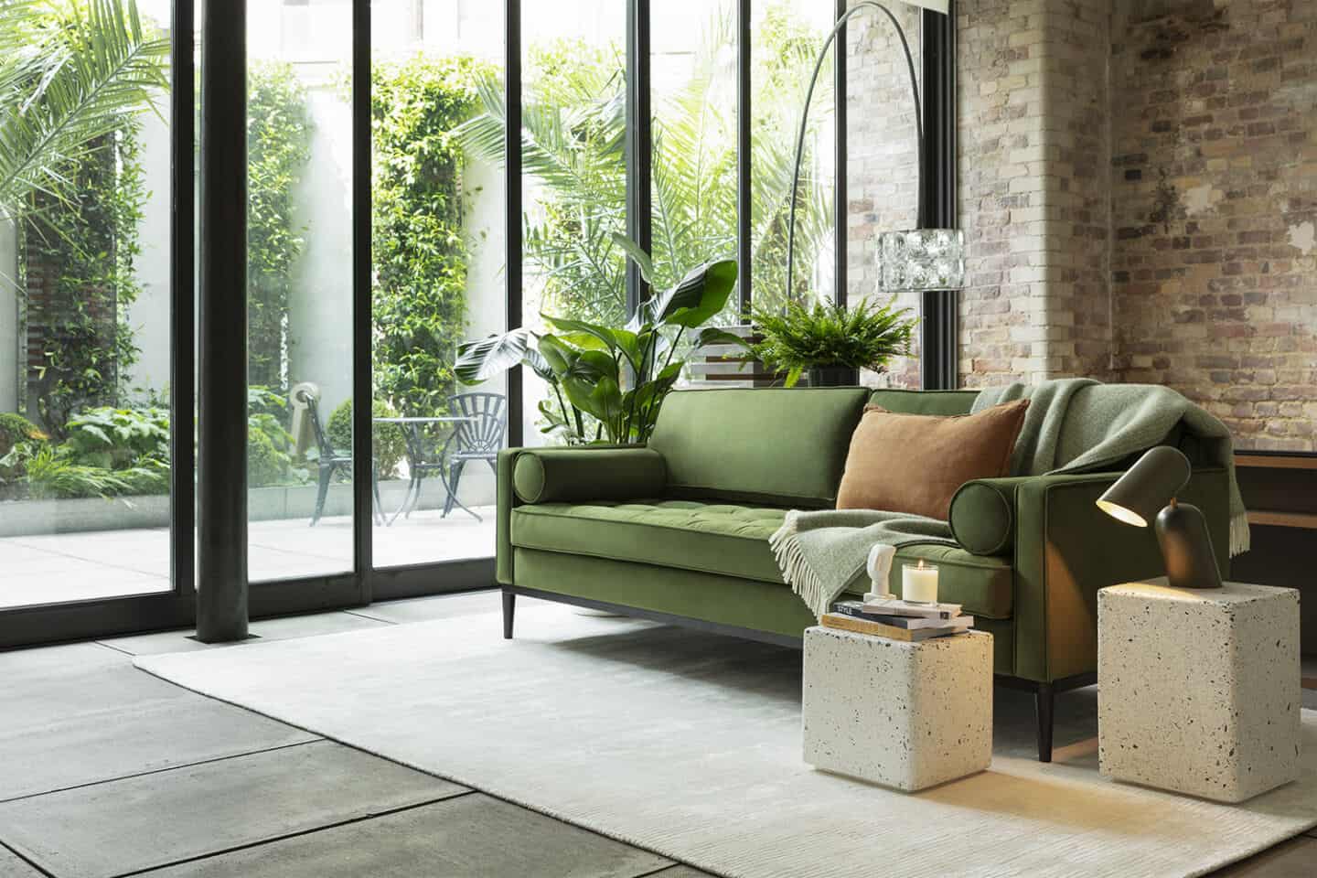 A green sofa from Swyft Home on a grey rug positioned next to a window with lots of plants outside and with a brick wall behind