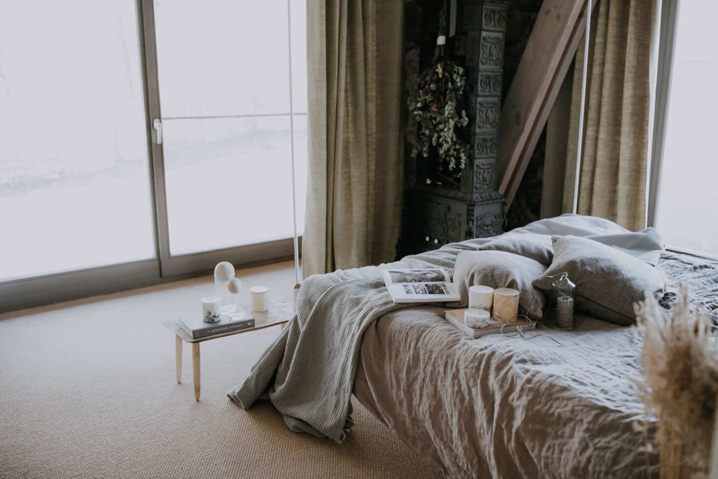 Textured Lives. Using texture in interiors to bring your home to life.  A bedroom with floor to ceiling wondows and natural light pouring in. The bed has a linen cover and there are books, candles and cushions on the bed.