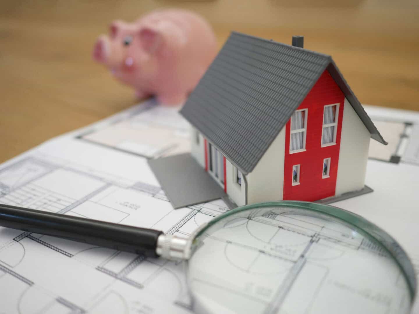 A magnifying glass and a small house on some architectural plans on a table with a piggy bank in the background.