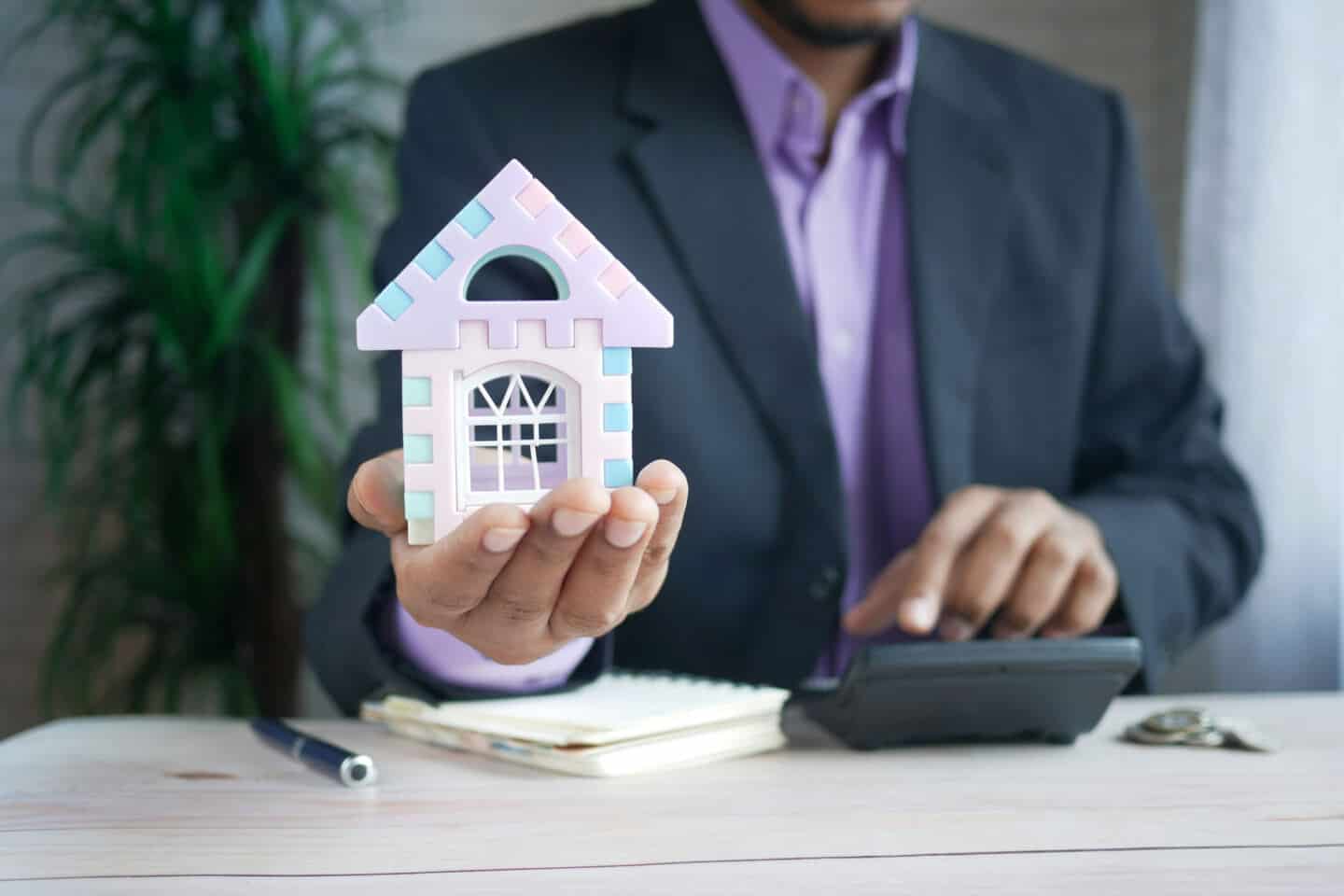 a man in a suit using a calculator holds a model house in his right hand