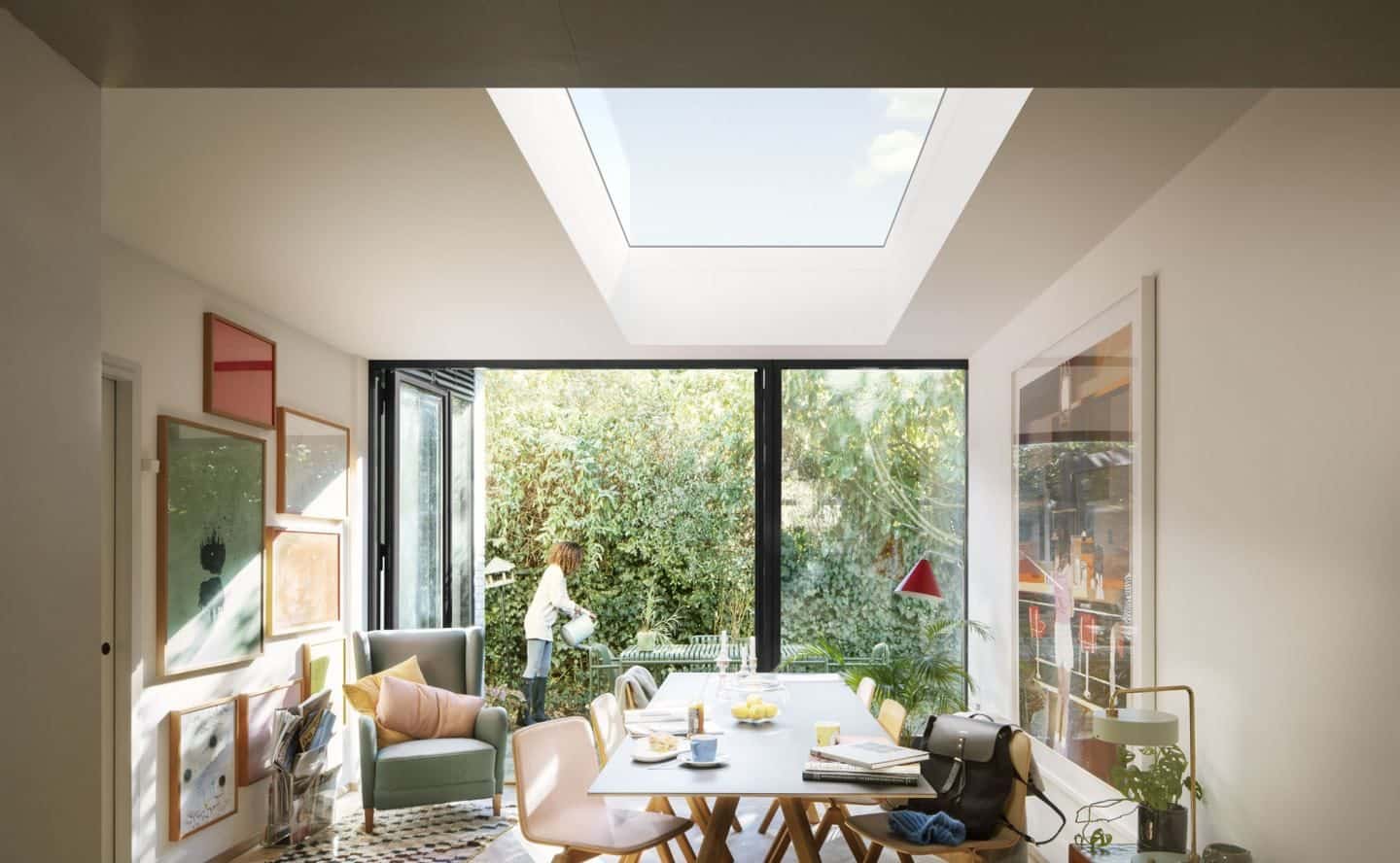 Vario by VELUX Bespoke Flat Roof Windows pictured above a dining room. Women seen outside bi-fold doors watering the garden.