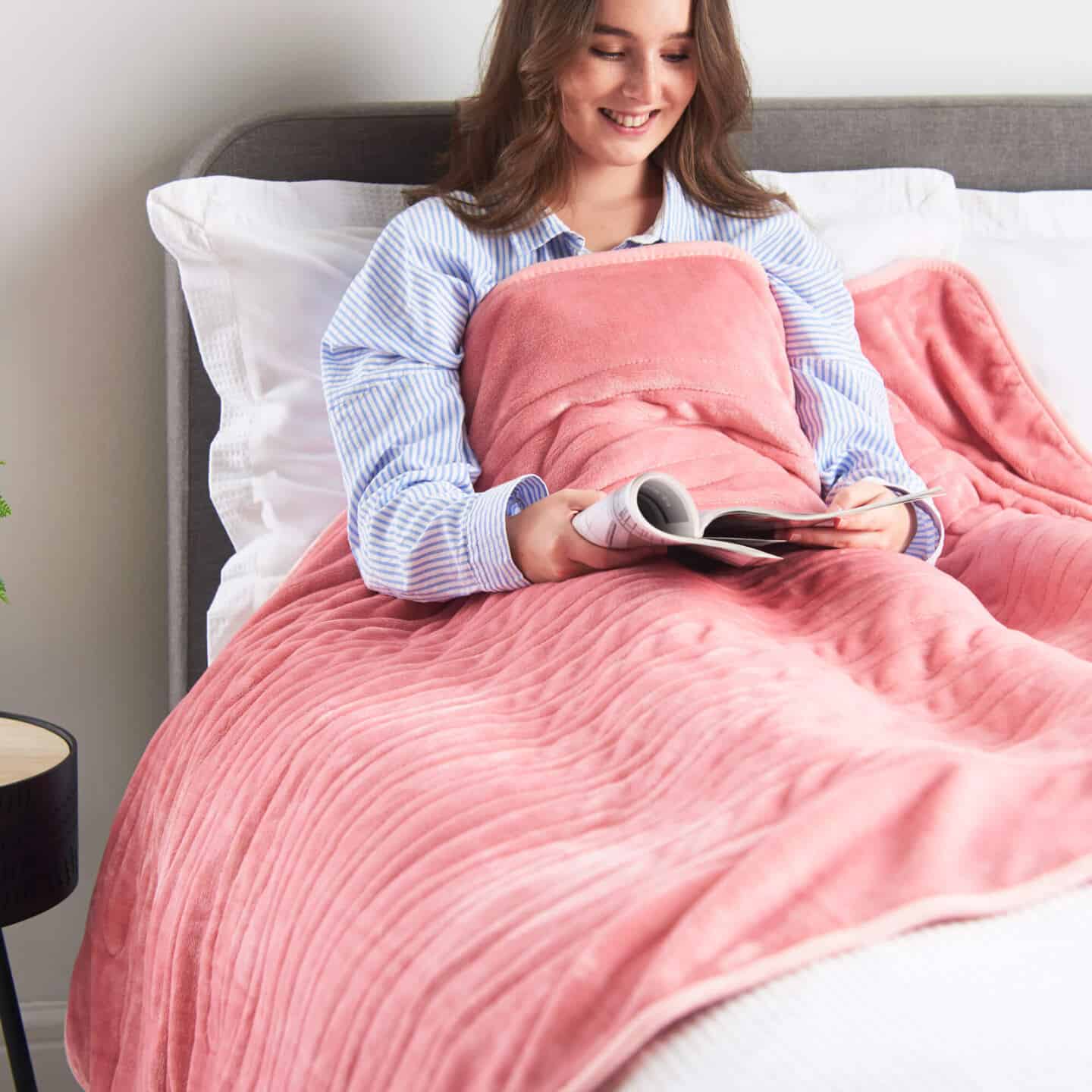 A young white woman in bed reading under a pink heated blanket