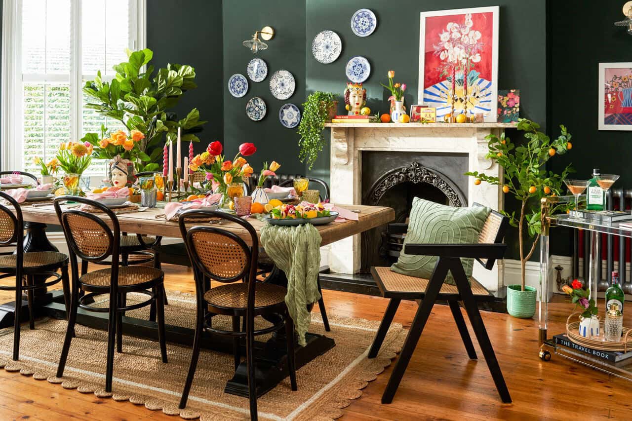 A dark grren dining room with colourful artwork and trinkets on the fireplace mantel. A chunky wooden table with rattan bistro chairs and lots of flowers adorn the table. A bar cart is the corner of the room