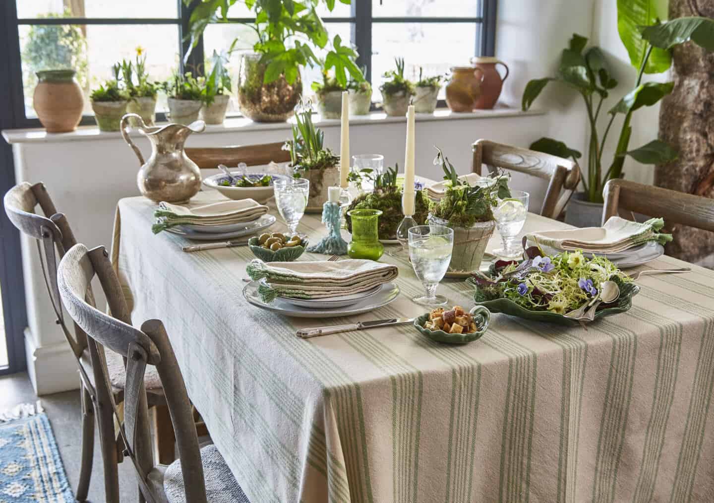 A dining room with table and four wooden chairs. Table is dressed with linen table cloth and lots of greenery. The window sill behind is covered in plants.