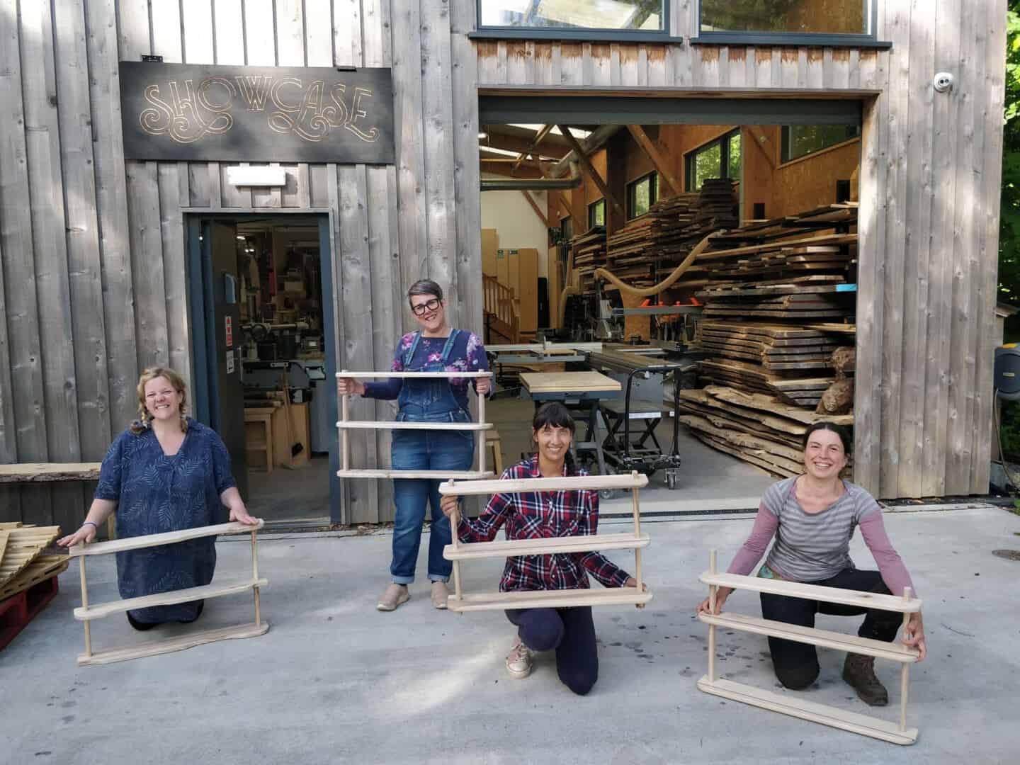 Four women stand outside of a workshop holding shelving units that they created on a woodwork course at The Woodlab iat Dartington in Devon 